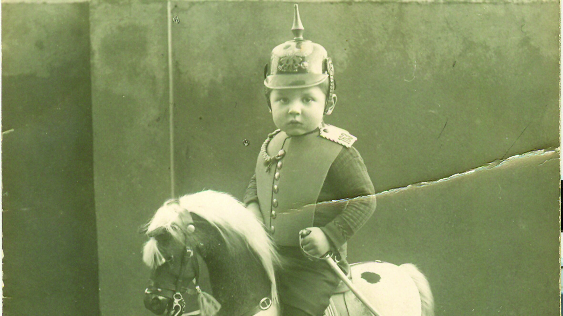 Black-and-white photo postcard of a boy, aged about three, wearing a toy uniform on a rocking horse. 