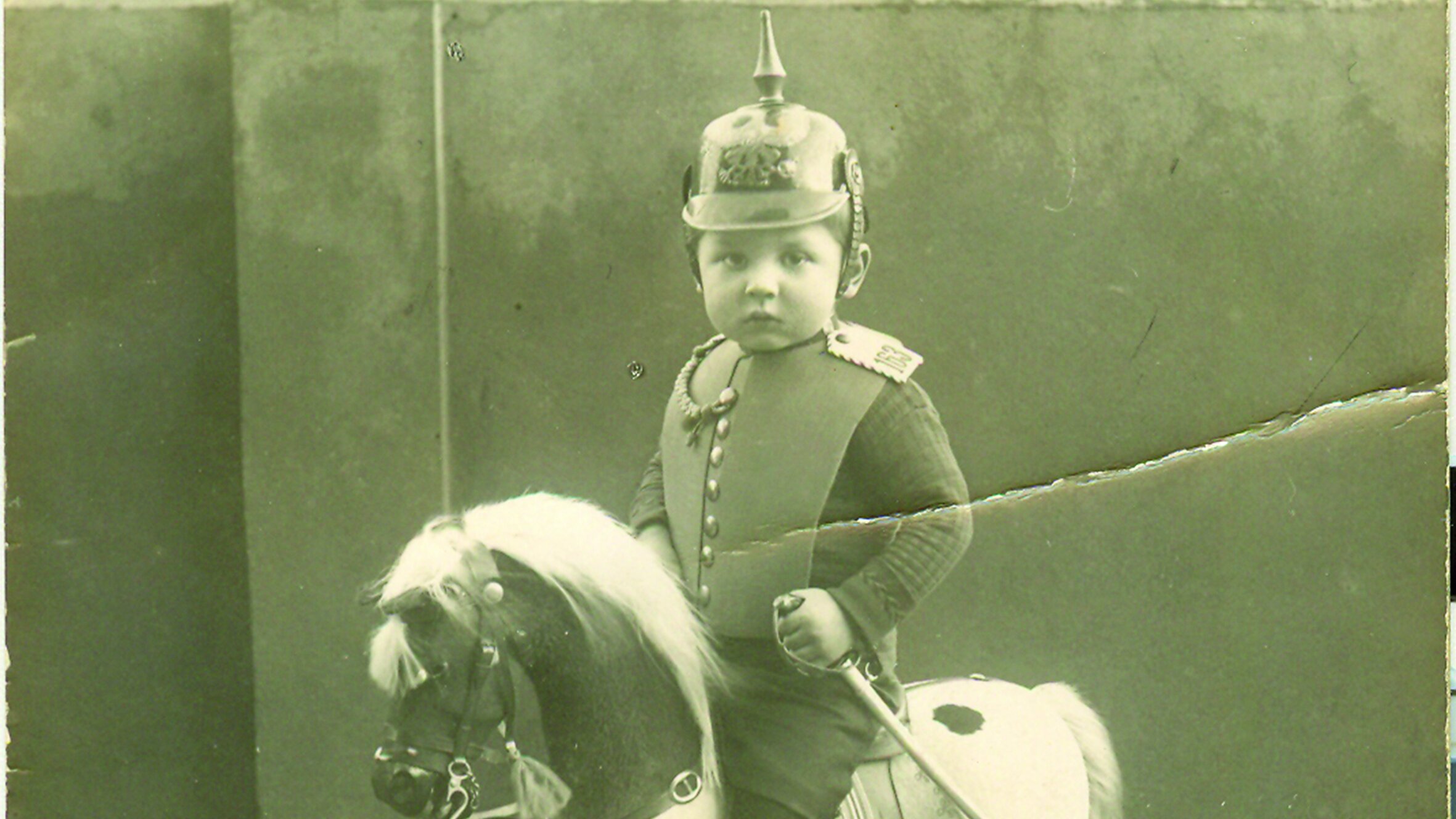 Die schwarz-weiße Fotopostkarte zeigt einen ca. dreijährigen Jungen in Spielzeuguniform auf einem Schaukelpferd. 
