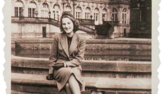 Das Schwarzweißfoto zeigt eine elegante junge Frau, die im Innenhof des Zwingers sitzend lachend in die Kamera schaut. 
