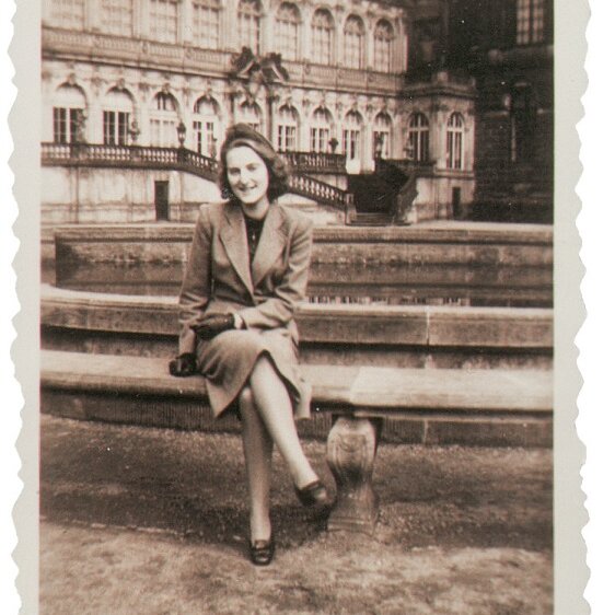 Das Schwarzweißfoto zeigt eine elegante junge Frau, die im Innenhof des Zwingers sitzend lachend in die Kamera schaut. 