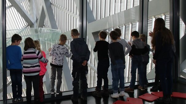 Ein weiblicher Guide und elf Kinder schauen im Ausstellungsbereich Dresden Blick aus einem großen Fenster auf die Stadt. 