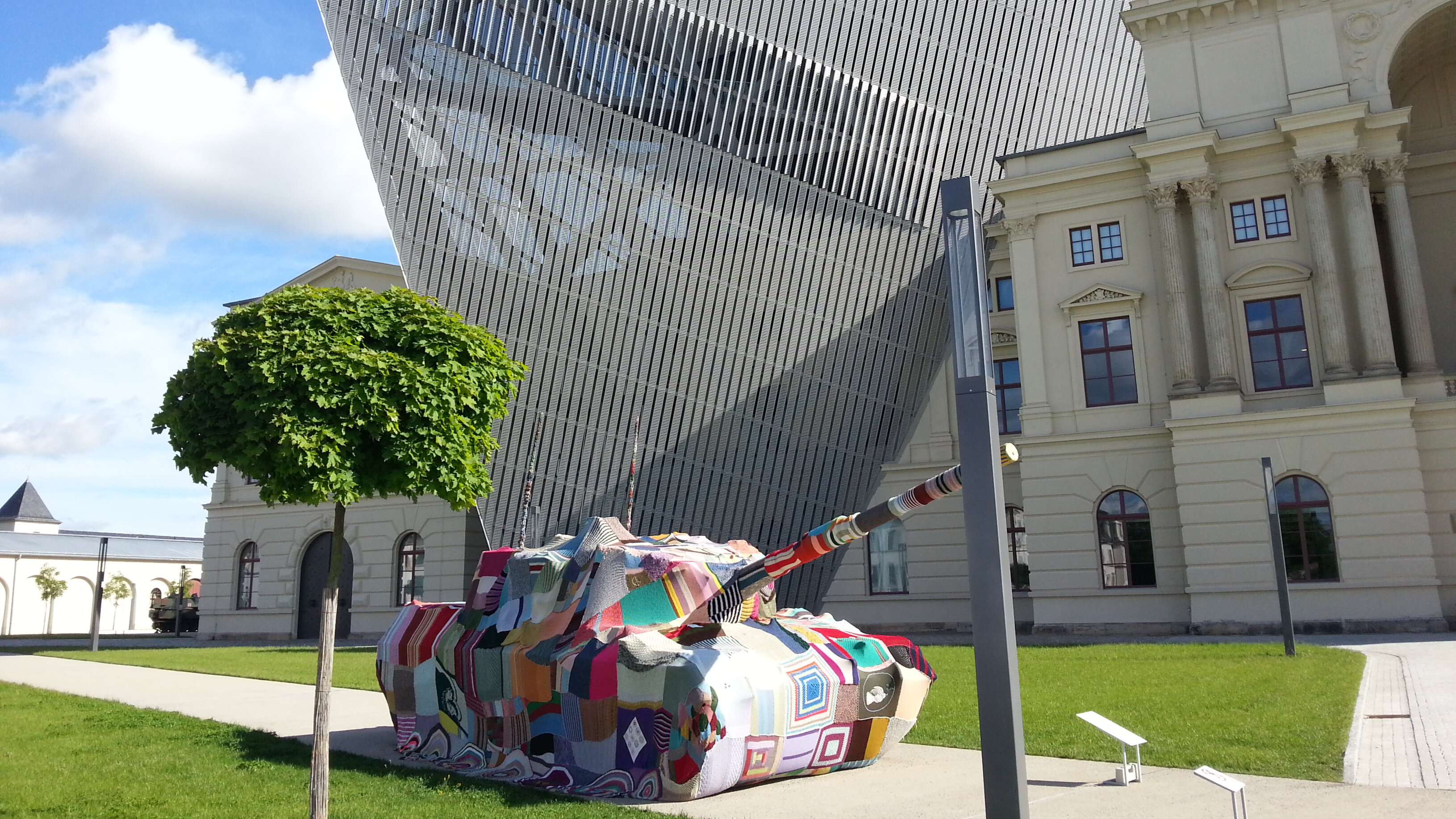 Das Bild zeigt einen Panzer, in einer gestrickten bunten Umhüllung, der vor dem MHM steht.  