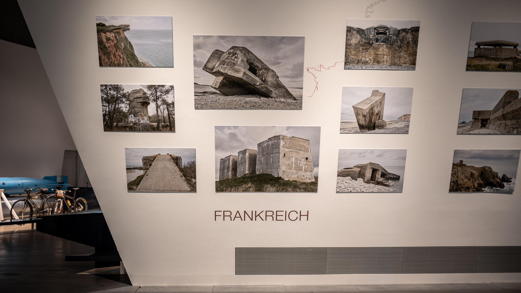 Zu sehen ist eine schräge Wand, auf der Bilder von Bunkeranlagen in Frankreich zu sehen sind.