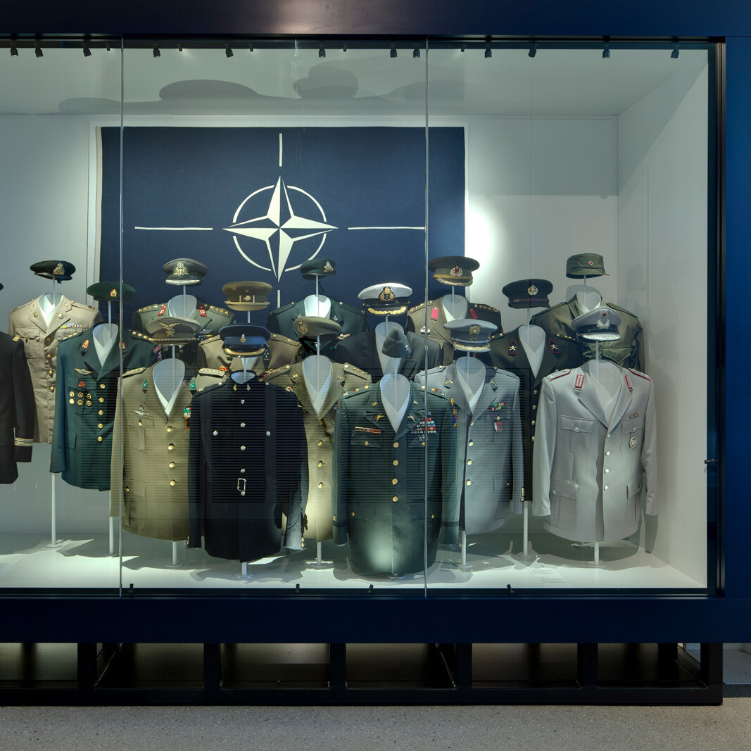 Das Bild zeigt eine Vitrine zur NATO in der Uniformen unterschiedlicher Mitgliedsländer hängen. 