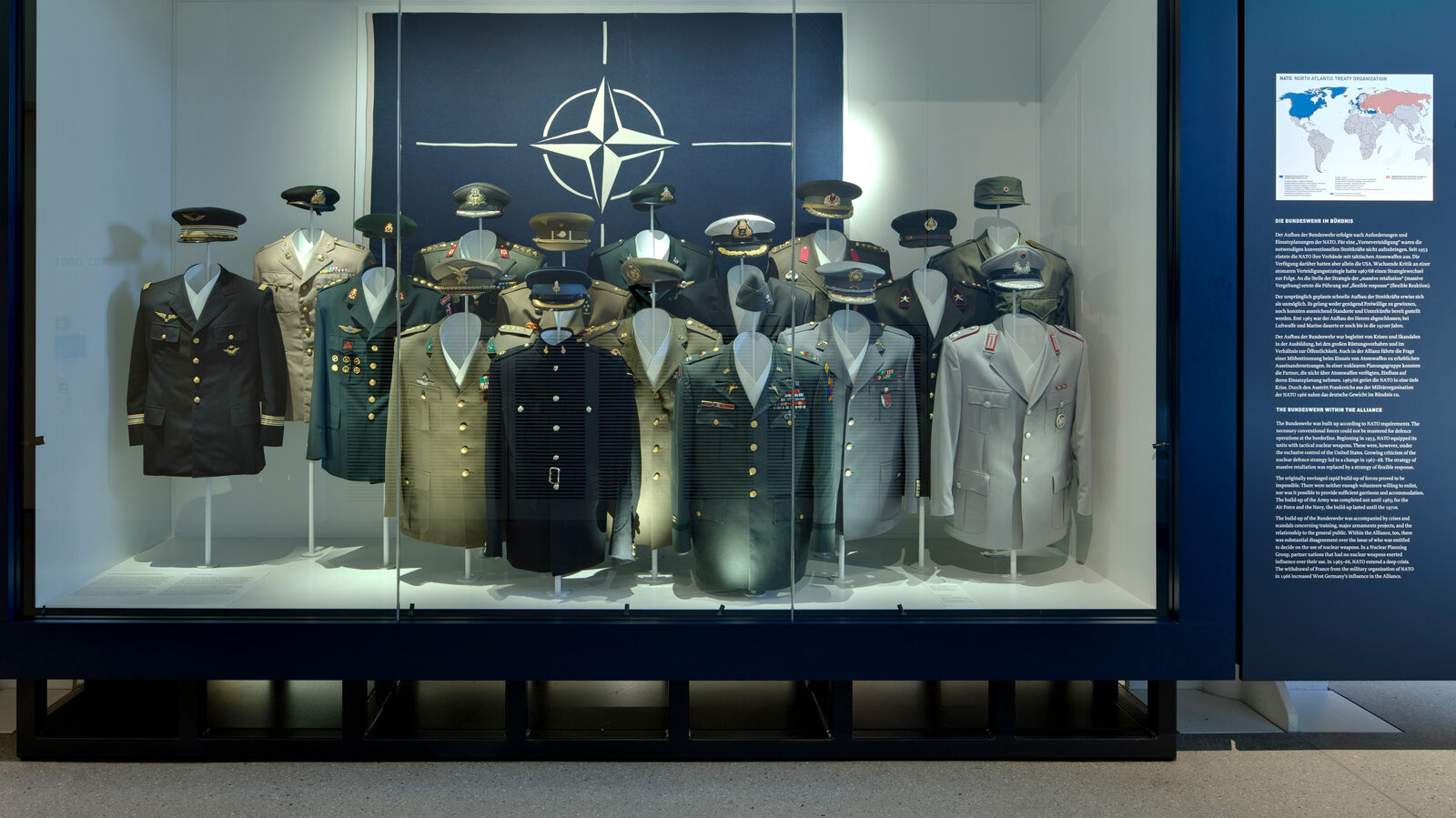 Das Bild zeigt eine Vitrine zur NATO in der Uniformen unterschiedlicher Mitgliedsländer hängen. 