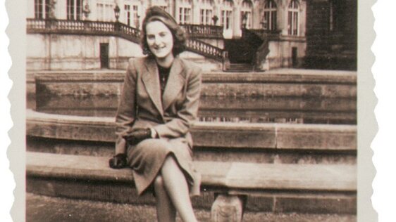 Das Schwarzweißfoto zeigt eine elegante junge Frau, die im Innenhof des Zwingers sitzend, lachend in die Kamera schaut. 